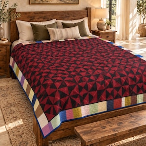 May include: A quilt featuring a repeating geometric pattern of dark red and black triangles. The quilt has a border composed of alternating rectangular blocks in shades of blue, green, yellow, brown, and white, finished with a solid blue edge.