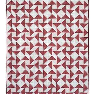 May include: A rectangular quilt with a repeating pattern of red and white geometric shapes. The design features alternating red triangles and white squares, creating a star-like motif. The quilt is likely made of fabric and has a stitched border.