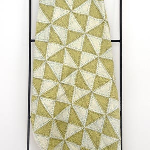 May include: A geometric patterned quilt in shades of olive green and cream, displayed on a black metal rack. The quilt features a repeating triangle design with visible stitching, creating a textured appearance. The overall aesthetic is modern and minimalist.