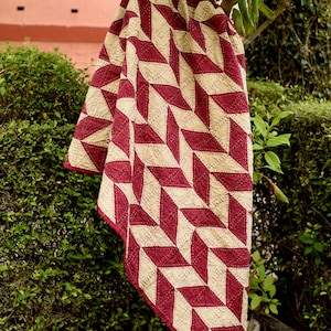 Può includere: Una trapunta piegata con un motivo geometrico a forma di rombi bordeaux e color crema. La trapunta è drappeggiata su un ramo d'albero, con un cespuglio verde e un muro rosa sullo sfondo. I bordi della trapunta sono rifiniti con tessuto bordeaux.