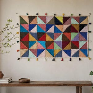May include: A rectangular wall hanging featuring a patchwork of multicolored fabric triangles in shades of red, blue, green, yellow, and pink. The piece is finished with a cream-colored border and small, dangling tassels along all four edges.