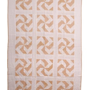 May include: A rectangular quilt featuring a repeating geometric pattern of white and tan pinwheel shapes arranged in a grid. The quilt is bordered by a solid tan edge and displays visible white stitching throughout the fabric surface.
