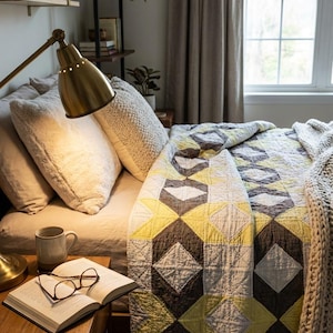 May include: A bedroom scene featuring a bed with a geometric patterned quilt in shades of yellow, gray, and brown. A brass lamp illuminates the bed, nightstand, and open book with eyeglasses. Decorative pillows and a mug are also visible.
