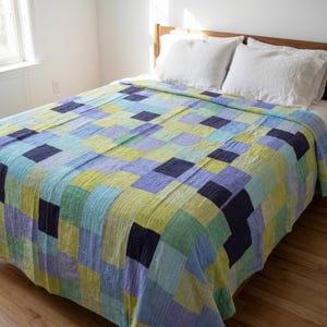 May include: A patchwork quilt in blue, green, and yellow hues covers a bed. The quilt has a geometric pattern of squares and rectangles. Two white pillows are on the bed. The quilt is made of a soft, textured fabric. The bed is made of wood.