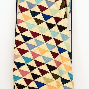 Op de afbeelding: Een kleurrijk quilt met een geometrisch driehoekpatroon. De quilt heeft driehoeken in verschillende kleuren, waaronder rood, blauw, geel en groen, gestikt op een crèmekleurige achtergrond. De quilt hangt aan een zwart metalen rek.
