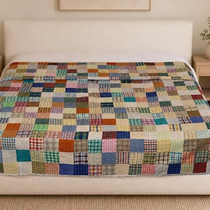 May include: A patchwork quilt featuring a grid of small, multi-coloured fabric squares in various patterns including plaid, gingham, and solid colours. The quilt is spread across a bed, displaying a diverse array of hues such as blue, red, green, yellow, and brown, creating a textured, multi-patterned surface that covers the entire mattress area.