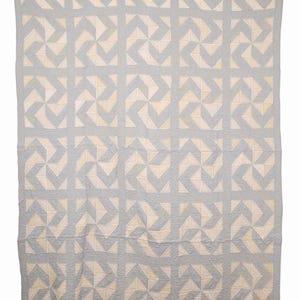 Puede incluir: Una colcha rectangular con un patrón geométrico repetitivo de cuadrados de tela azul claro y blanco roto. El diseño consiste en formas de molinillo de cuatro puntas dispuestas en una cuadrícula, con costuras visibles en la superficie.