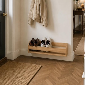 May include: A light-toned wooden wall-mounted coat rack with six black metal hooks and a top shelf, holding a beige jacket and a small potted plant. Below it, a matching light-toned wooden wall-mounted shoe rack holds two pairs of shoes.