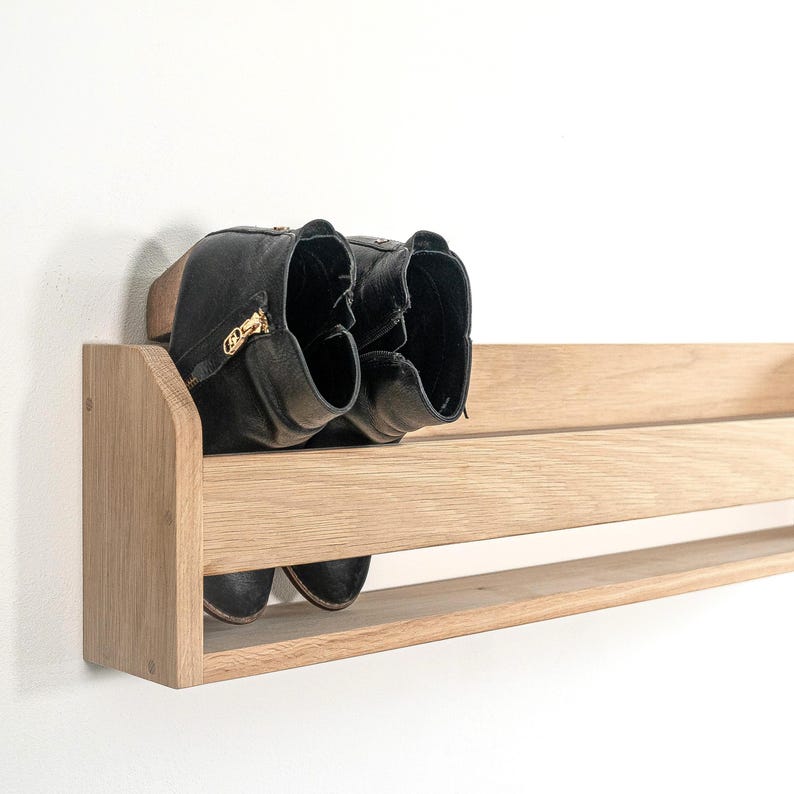 May include: A wooden shoe rack mounted on a white wall. The rack has two shelves and is made of light brown wood. Two black boots are on the top shelf.