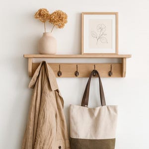 May include: A light oak wall-mounted shelf with four bronze hooks. On the shelf sits a beige ceramic vase with dried flowers and a framed line drawing of a flower. A tan trench coat and a two-toned beige and olive green canvas tote bag hang below.