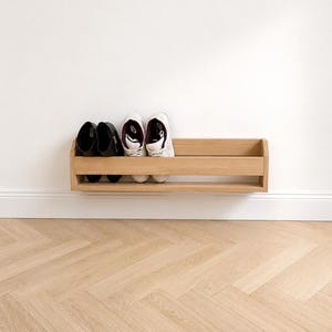 Pode incluir: Um sapateiro de parede em madeira clara com um design de prateleira horizontal. O suporte cont&eacute;m um par de botins pretos e um par de t&eacute;nis brancos, oferecendo uma solu&ccedil;&atilde;o de arruma&ccedil;&atilde;o para cal&ccedil;ado num ambiente dom&eacute;stico.