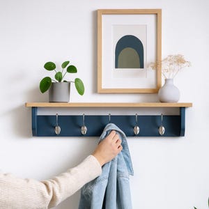 May include: A navy blue and natural wood wall shelf with five silver hooks. A framed print with an abstract design is above the shelf, and a small potted plant and vase with dried flowers are displayed. A denim jacket hangs from a hook.