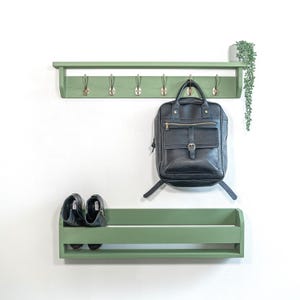 Op de afbeelding: Een groene houten wandplank met een plank en haken. Een zwarte leren rugzak hangt aan een van de haken. Twee paar zwarte schoenen staan op de plank onder de haken.