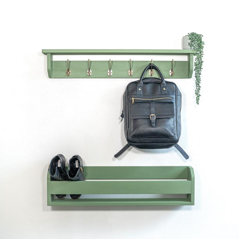 Op de afbeelding: Een groene houten wandplank met een plank en haken. Een zwarte leren rugzak hangt aan een van de haken. Twee paar zwarte schoenen staan op de plank onder de haken.