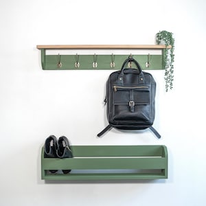 May include: A green wall-mounted coat rack with a shelf and a shoe shelf. The rack has six hooks and a black backpack hanging from one of the hooks. There are two pairs of black shoes on the shoe shelf.