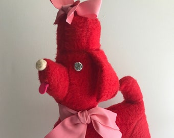 May include: Red plush toy dog with pink bows. The dog has a button nose and a sparkly button on its chest.