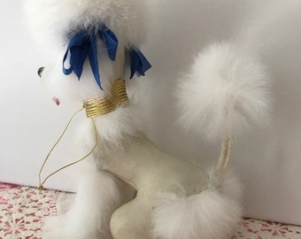 May include: White plush poodle ornament with blue bows and a gold collar. The poodle has a fluffy tail and is sitting on a pink and white doily.