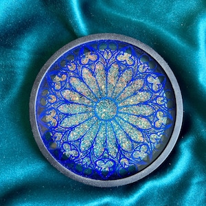 Peut inclure: Un dessous de verre circulaire présentant un motif de rosace gothique bleu foncé superposé à une base remplie de paillettes irisées multicolores. Le design est enchâssé dans un cadre circulaire noir mat lisse, créant un contraste net.