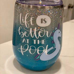 May include: A stemless wine tumbler with a glittery silver and blue ombre design. The tumbler has the phrase "life is better at the pool" and a cartoon unicorn floatie. The tumbler has a clear lid.