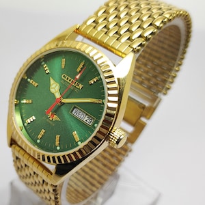 Può includere: Un orologio automatico Citizen color oro con quadrante verde. L'orologio presenta lancette, indici e un display giorno-data dorati. Il cinturino dell'orologio è in tinta oro.