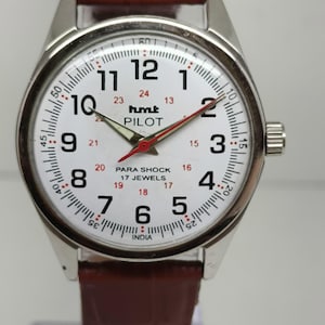 May include: A silver wristwatch with a white face, black numerals, and a red second hand. The watch has the text "hmt PILOT" and a brown leather strap. The watch face displays the time and has a 17 jewels marking.