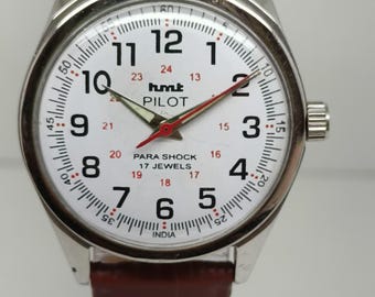 Puede incluir: Reloj de pulsera plateado con esfera blanca, números negros y segundero rojo. El reloj presenta el texto "hmt PILOT" y una correa de cuero marrón. La esfera muestra la hora y tiene la marca 17 jewels.