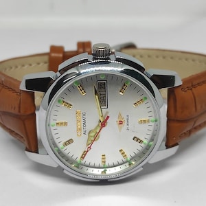 Op de afbeelding: Een zilverkleurig Citizen automatisch horloge met een lichtgekleurde wijzerplaat, gouden accenten en een bruine leren band. De wijzerplaat toont de dag en datum. De band heeft een textuur met krokodillenpatroon.