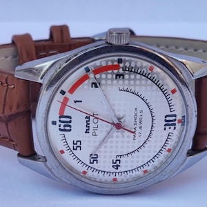 May include: A silver wristwatch with a white face and a brown leather strap. The watch face has black numerals, red details, and the text "hmt PILOT PARA SHOCK 17 JEWELS". The strap is brown.
