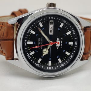 May include: A Citizen automatic wristwatch with a black dial, silver case, and brown leather strap. The watch face displays the day and date, with white numerals and markers. The watch has red and silver hands.