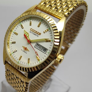 Puede incluir: Reloj Citizen Automatic dorado con esfera blanca, visualización de día y fecha y segundero rojo. Tiene un bisel texturizado y una correa de malla dorada. La esfera muestra "Citizen Automatic" y "21 Jewels".