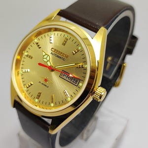 Pode incluir: Um relógio Citizen Automatic dourado com uma bracelete de couro castanho. O mostrador do relógio é dourado com marcadores pretos e verdes, um ponteiro de segundos vermelho e um visor de dia/data. O relógio é exibido em um suporte transparente.