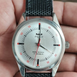 May include: Silver wristwatch with a black fabric strap. The watch face is silver with black and red markers, displaying the text "hmt PILOT" and "PARA SHOCK 17 JEWELS". The watch has a round face and a textured strap.