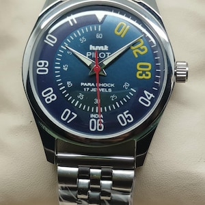 May include: A stainless steel wristwatch with a blue face and a silver strap. The watch face has yellow numerals, white markers, and the text "hmt PILOT". The watch also has a red second hand and the text "PARA SHOCK 17 JEWELS INDIA".