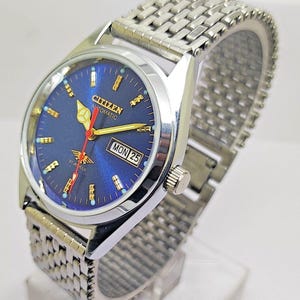 May include: A Citizen automatic wristwatch with a blue dial and a silver-tone stainless steel band. The watch face displays the day and date, with gold-tone hands and markers. The watch is a classic design.