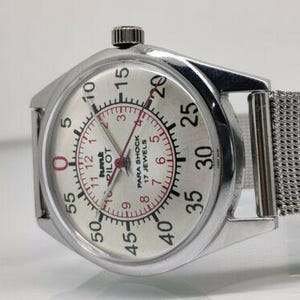 May include: A silver wristwatch with a mesh strap. The watch face is white with black numerals and red detailing. The watch reads "PILOT PARA SHOCK 17 JEWELS". The watch has a round face with a silver bezel.