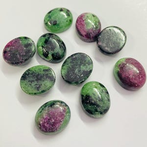 May include: A collection of oval-shaped zoisite gemstones with a mix of green and magenta colors. The polished stones have a glossy finish and are arranged on a white surface. These gemstones are suitable for jewelry making.