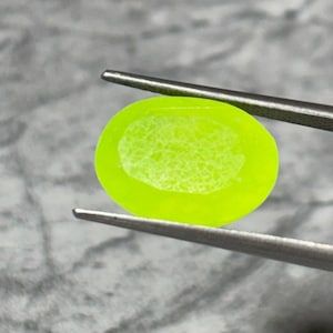 Puede incluir: Una gema ovalada de color verde neón es sostenida por unas pinzas. La gema tiene un patrón más claro, similar a una telaraña, en el centro. Las pinzas son plateadas y el fondo es gris borroso.