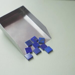 Puede incluir: Una cuchara de acero inoxidable contiene varias baldosas cuadradas de lapislázuli azul oscuro. Las baldosas tienen motas de oro y están dispersas sobre una superficie verde claro. La cuchara está en ángulo y las baldosas están en primer plano.