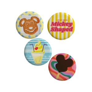 May include: Four circular pin-back badges featuring snack-themed designs. One shows a Mickey-shaped pretzel on a blue polka dot background, another has yellow stripes with the text I LIKE MY SNACKS Mickey Shaped, a third displays a yellow soft serve treat, and the fourth features a chocolate-covered Mickey-shaped ice cream bar on a colourful wavy background.