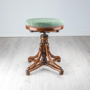 May include: A round stool with a sage green velvet upholstered seat and a dark brown wooden base. The stool features a central adjustable screw mechanism and four curved wooden legs that terminate in rounded feet, providing a stable structure.
