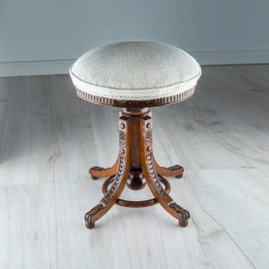 Puede incluir: Un taburete redondo con asiento de tela texturizada gris claro y base de madera marrón oscuro. La base cuenta con cuatro patas curvas con detalles florales tallados y un anillo de soporte central circular para la estabilidad.