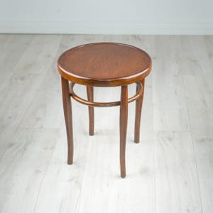 May include: A round wooden stool with a dark brown finish, featuring a circular seat and four curved legs connected by a circular support ring underneath the seat. The stool has a smooth, polished surface and a simple, functional design.