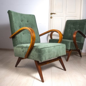 May include: Two green upholstered armchairs with wooden frames. The chairs have a curved back and arms, and the upholstery is a soft, textured fabric. The chairs are sitting on a light wood floor.