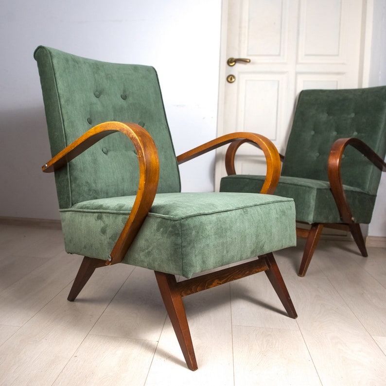 May include: Two green upholstered armchairs with wooden frames. The chairs have a curved back and arms, and the upholstery is a soft, textured fabric. The chairs are sitting on a light wood floor.