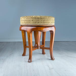 May include: A vintage wooden stool with a woven, patterned seat. The stool features curved legs and a rich, warm brown finish. The seat is a circular shape with a beige and brown checkered pattern.