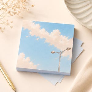 May include: A square notepad featuring a blue sky with fluffy white clouds and a street lamp illustration. The notepad is stacked with several sheets visible, and is resting on a light blue surface.