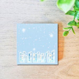 May include: A square, light blue sticky note with a grid pattern and a winter theme. The design features illustrated books at the bottom and snowflakes falling from the top. The books are in various colors and styles.