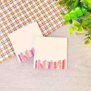 May include: Two square notepads with a row of colorful books printed at the bottom. The notepads are cream-colored with faint horizontal lines. A plaid orange and white patterned cloth is in the background.