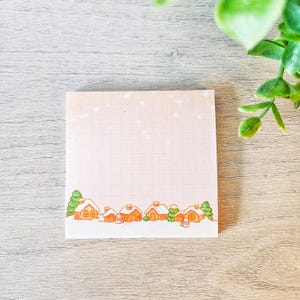 May include: Square sticky note with a winter village scene. The bottom of the note features a row of orange houses with snow-covered roofs and green trees. The background is a light pink with a grid pattern and white speckles.