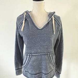 May include: A blue-grey hoodie with a deep V-neck and a kangaroo pocket. The hoodie has long sleeves and a drawstring hood. White stitching accents the edges of the pocket, hood, and sleeves. The garment is displayed on a mannequin.